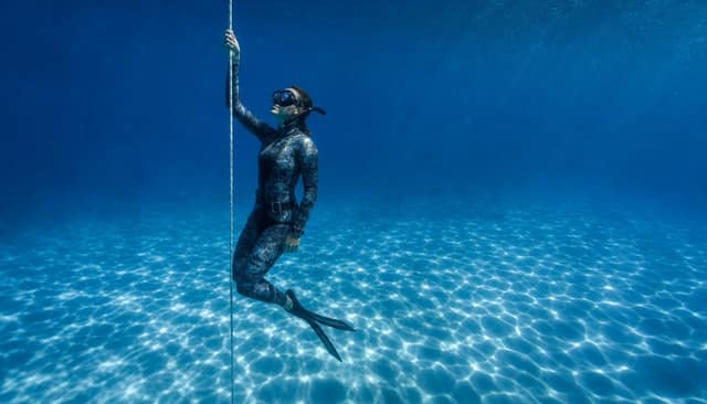 A freediver hanging on the line above a pale, rippled sandy seabed.