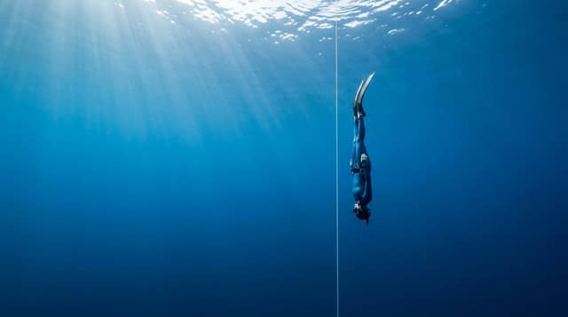 A freediver in freefall, head-down on the line as the light rays fan out above.
