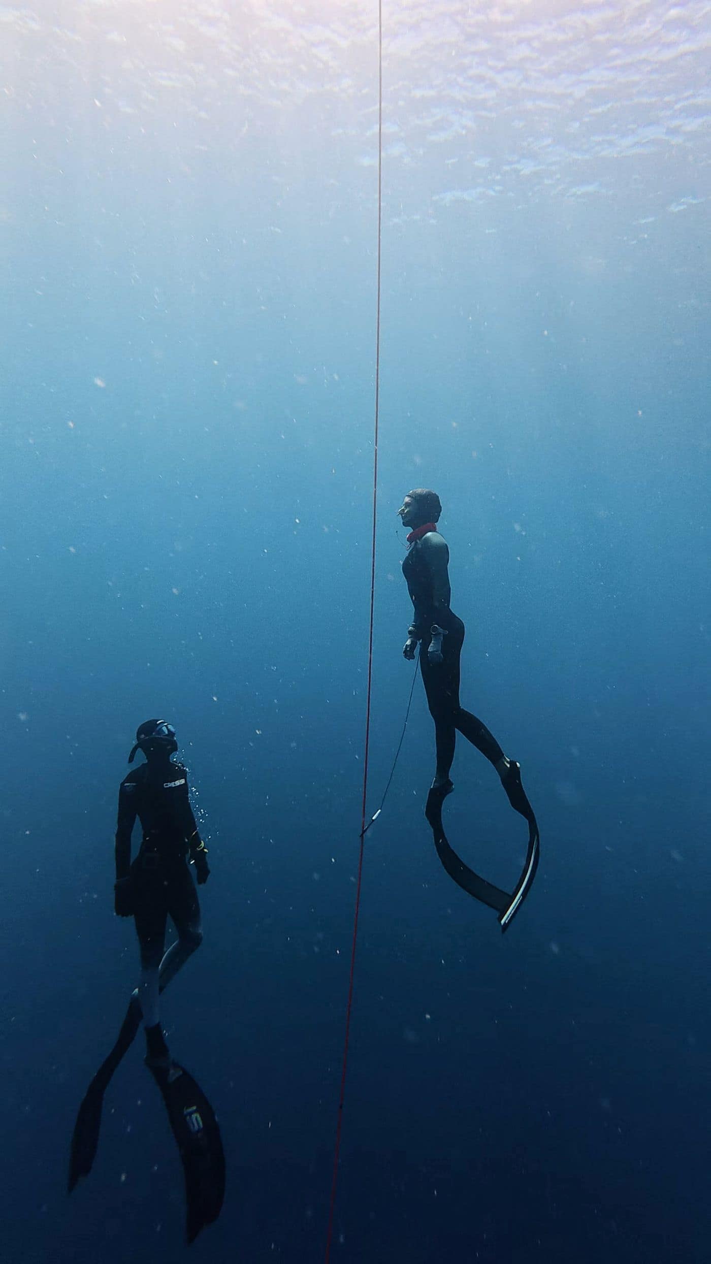 Two freedivers hanging weightless on the descent line in deep blue water, the bright surface far above.